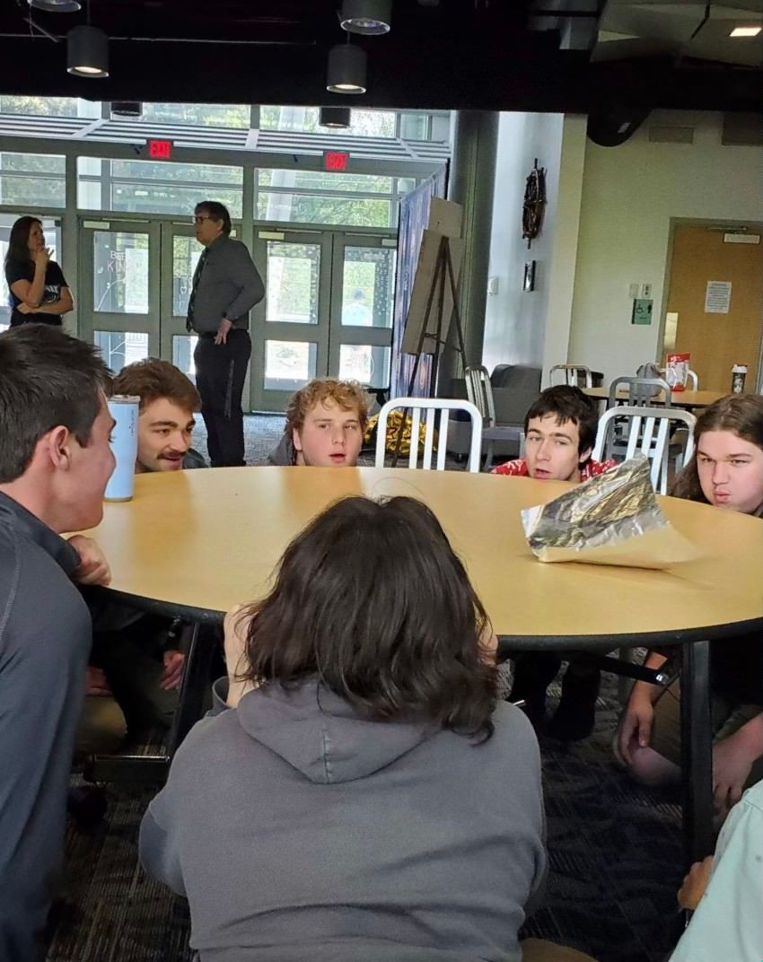 Students circle around a table and lift a piece of tin foil with their breath.