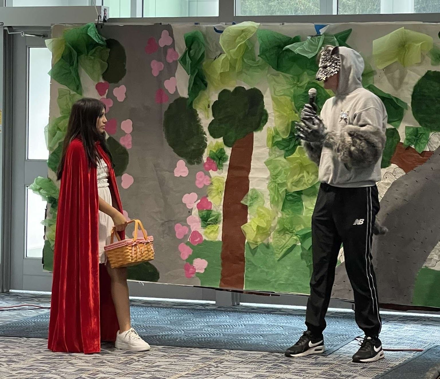 A girl in a red cloak stands looking at a person dressed in a wolf mask.